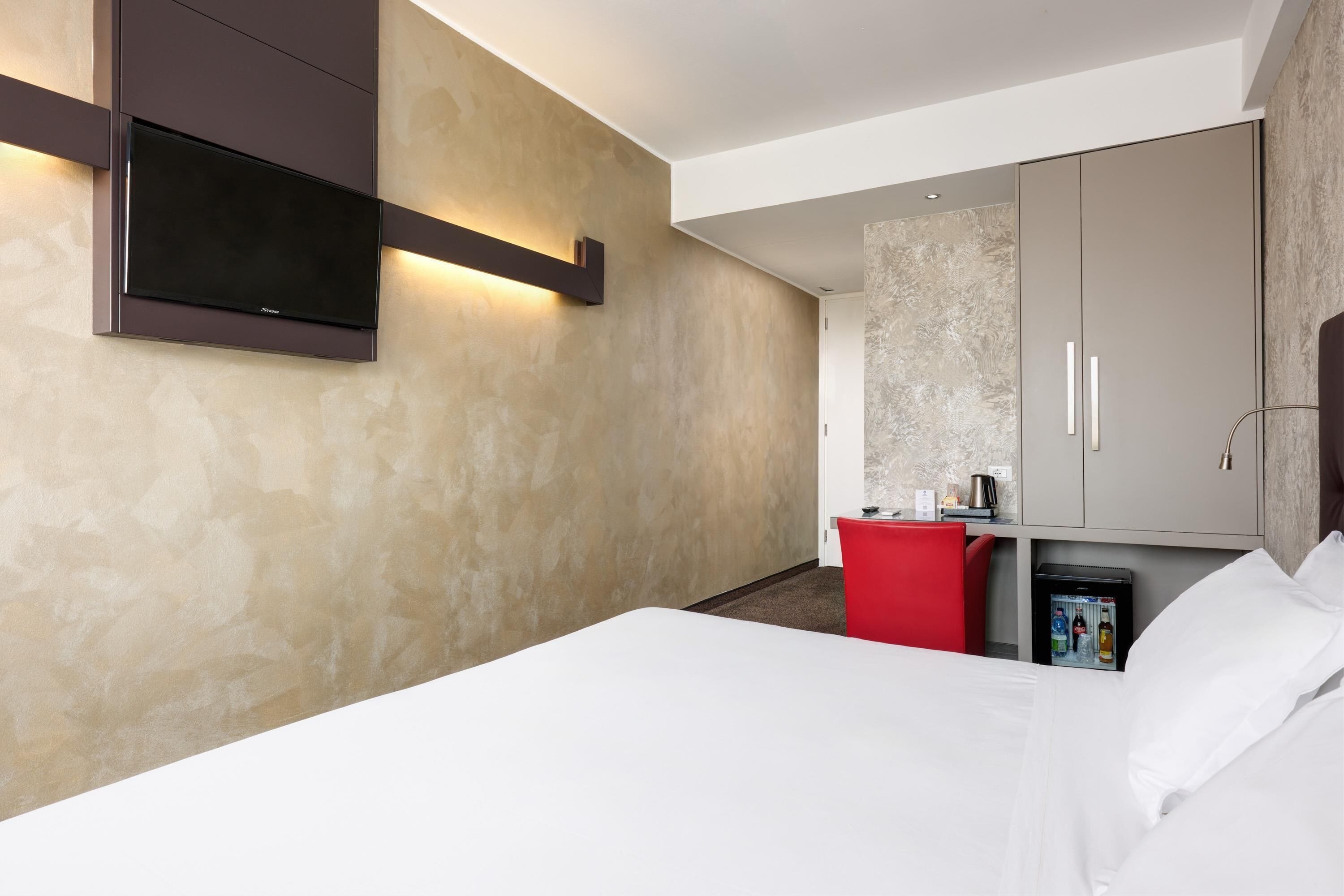 Une chambre d’hôtel avec un lit, un téléviseur fixé au mur, un minibar et un bureau rouge.