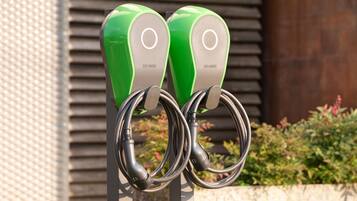 Zwei grüne Ladestationen für Elektroautos mit einer Metallsäule vor einem Gebäude mit horizontalem Verkleidungsmuster und etwas Grün.