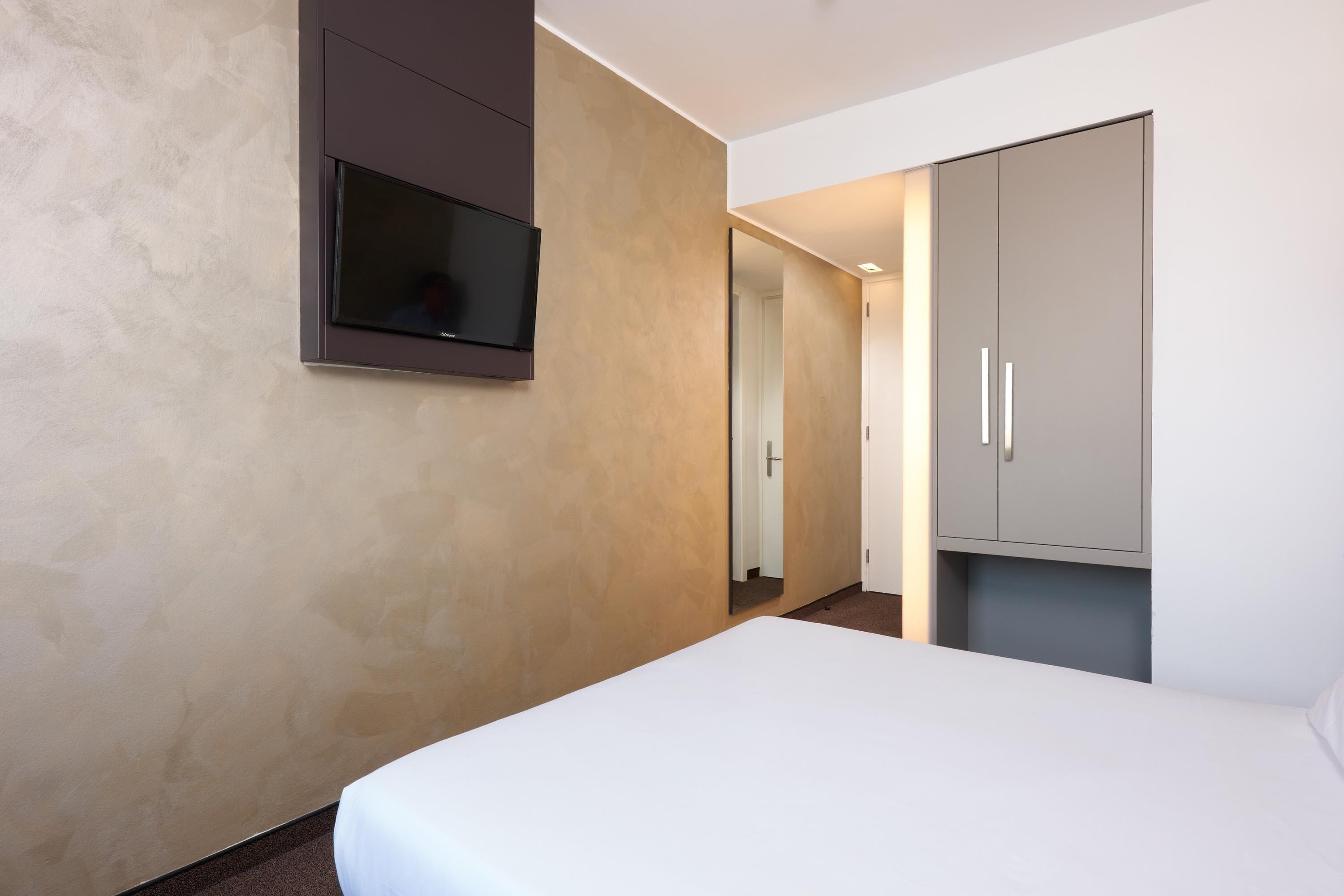 A hotel room with a bed, a television mounted on the wall, and a wardrobe.