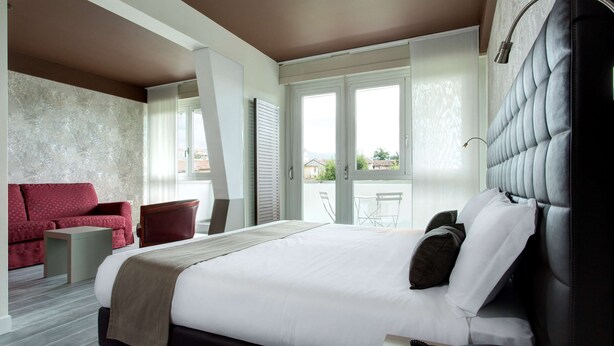 A modern hotel room with a large bed, a red sofa, a small table, and a view of the outdoors through large windows.