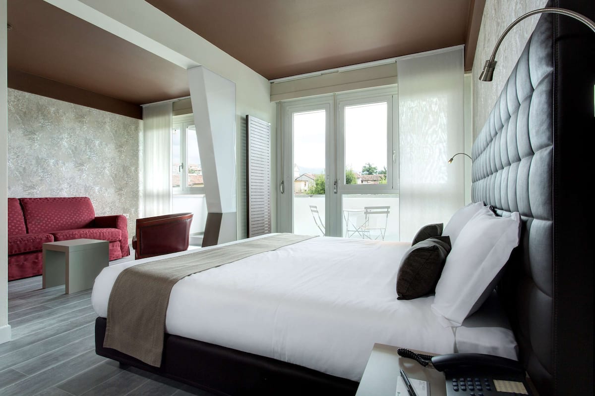 Una camera d'albergo moderna con un letto grande, un divano rosso, un tavolino e vista sull'esterno attraverso ampie finestre.