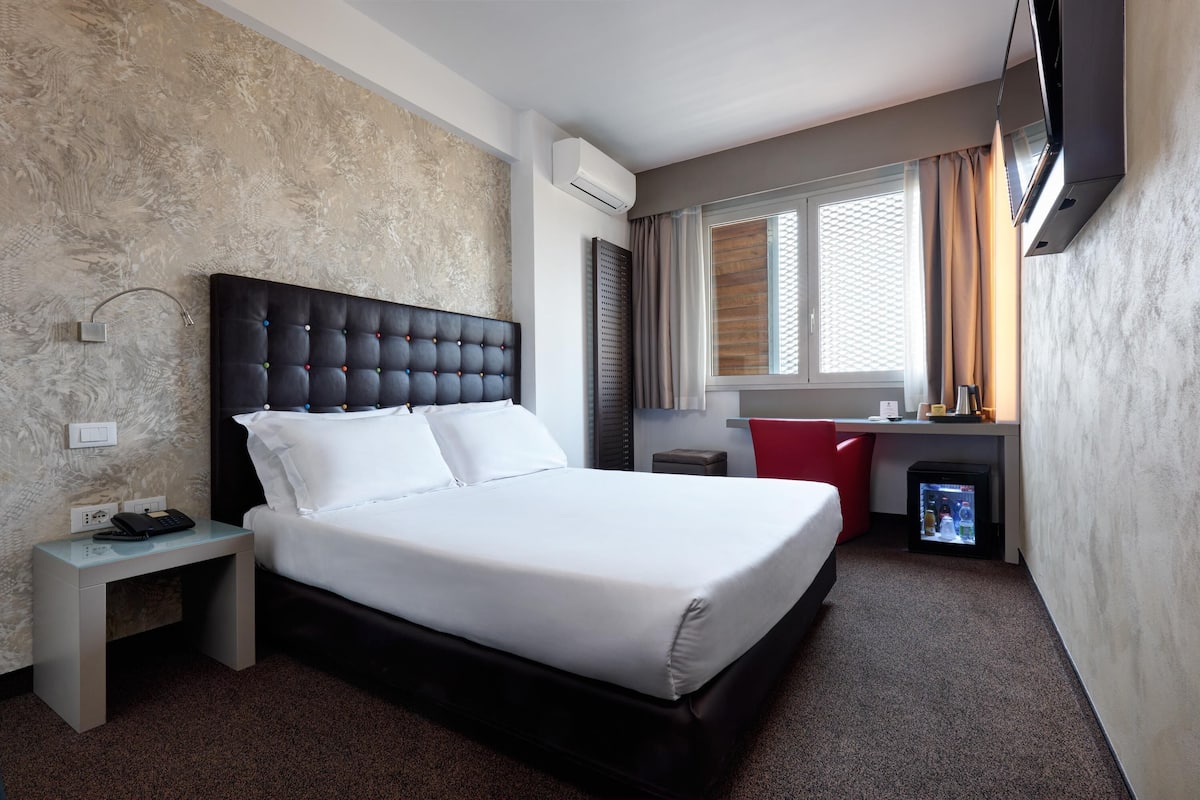 Una camera d'albergo con un letto, un comodino, una TV e una piccola area scrivania.