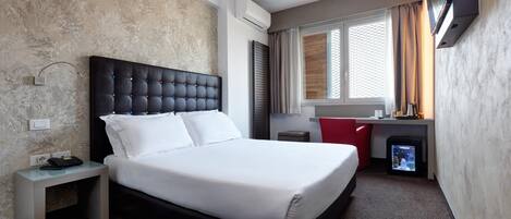 Una habitación de hotel con cama, mesita de noche, televisión y un pequeño escritorio.