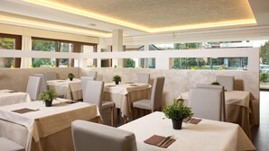 A modern restaurant with tables covered in beige tablecloths, chairs, and potted plants.