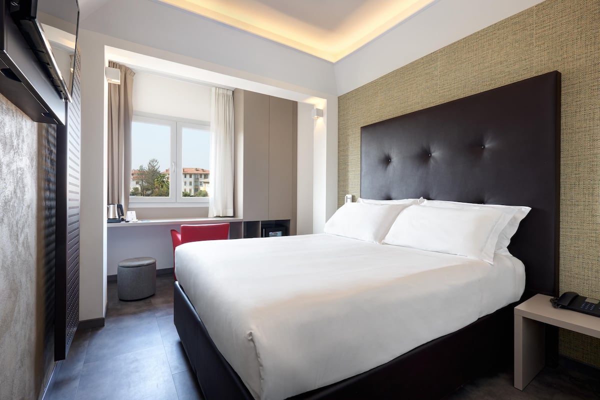 Una camera d'hotel moderna con un letto grande, un comodino con il telefono, una finestra con tende e vista sugli edifici.