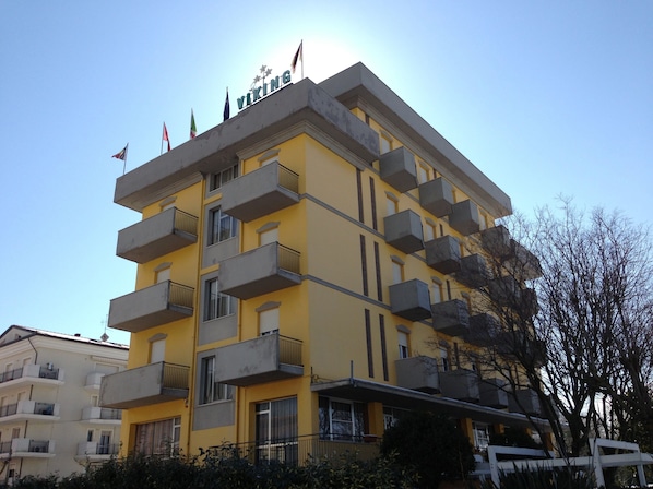 A multi-story yellow building with balconies, a clear sky, and flags on top. - Hotel Viking (Rimini)