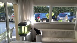 Ein Wohnzimmer mit einem weißen Sofa, einem Aquarium auf einem Tisch und einem Fenster mit Blick auf geparkte Autos und Grünpflanzen.