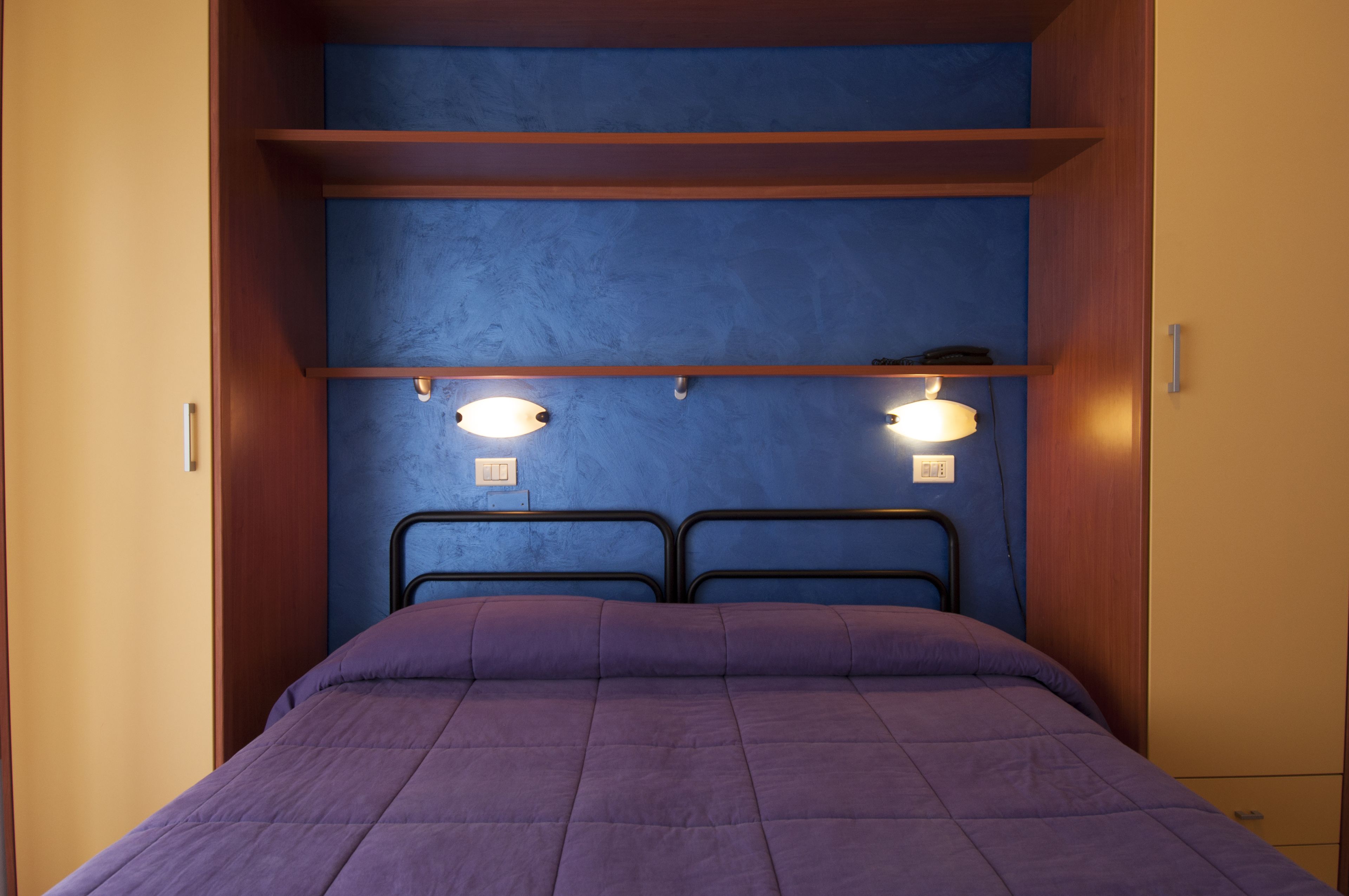 A single bed with a purple bedspread in a room with blue walls and wooden trim. There are two wall-mounted light fixtures and a shelf above the bed.