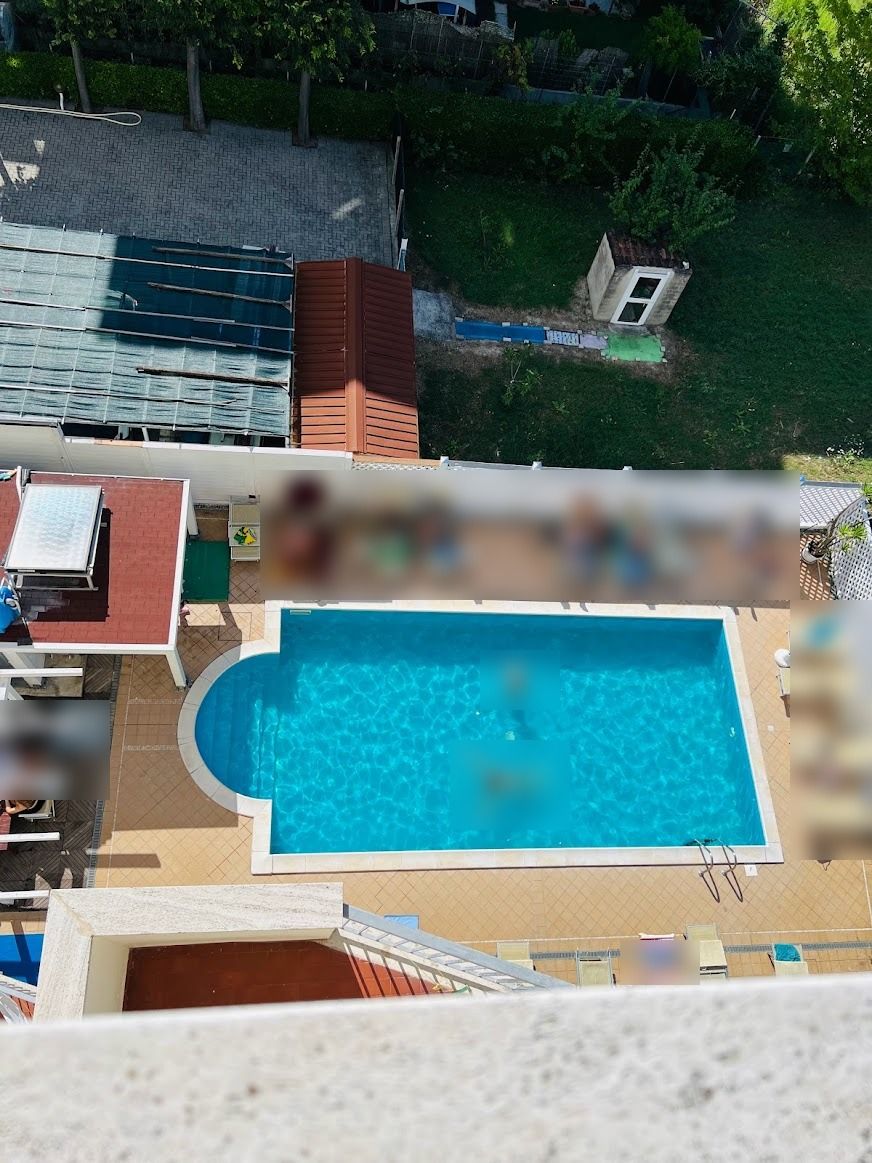 A swimming pool with a fenced deck area, surrounded by a residential setting with a small building and greenery.