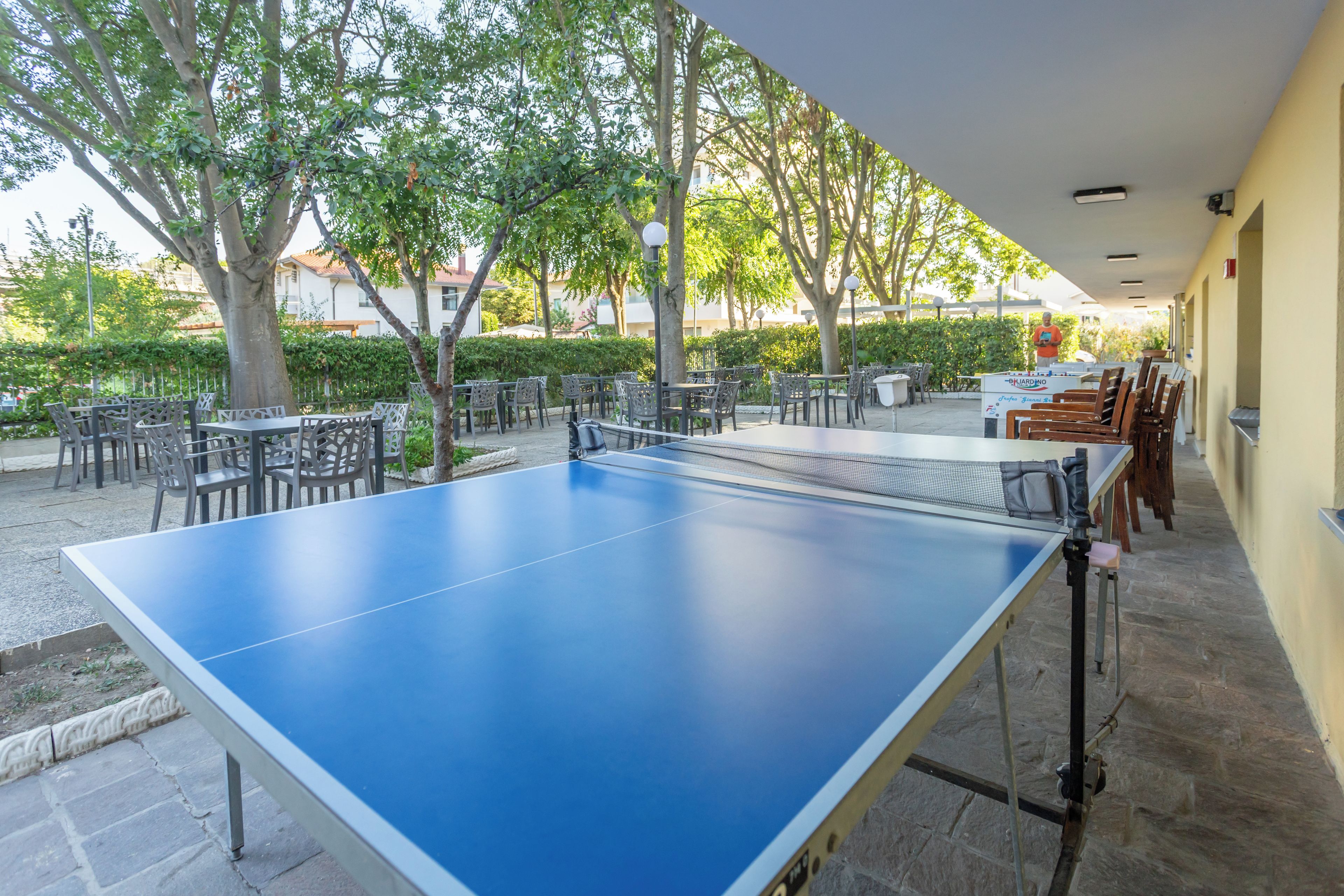 Una mesa de ping pong al aire libre con sillas alrededor, con árboles y un edificio como fondo.