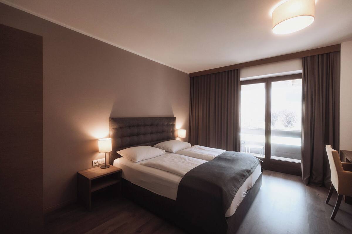 Une chambre d’hôtel avec un grand lit, deux lampes de chevet, une fenêtre avec des rideaux et un fauteuil.
