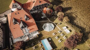 An aerial view of a large building with a red-tiled roof, multiple chimneys, and a swimming pool surrounded by lounge chairs. - Hotel Montis - Schönblick (Brunico)