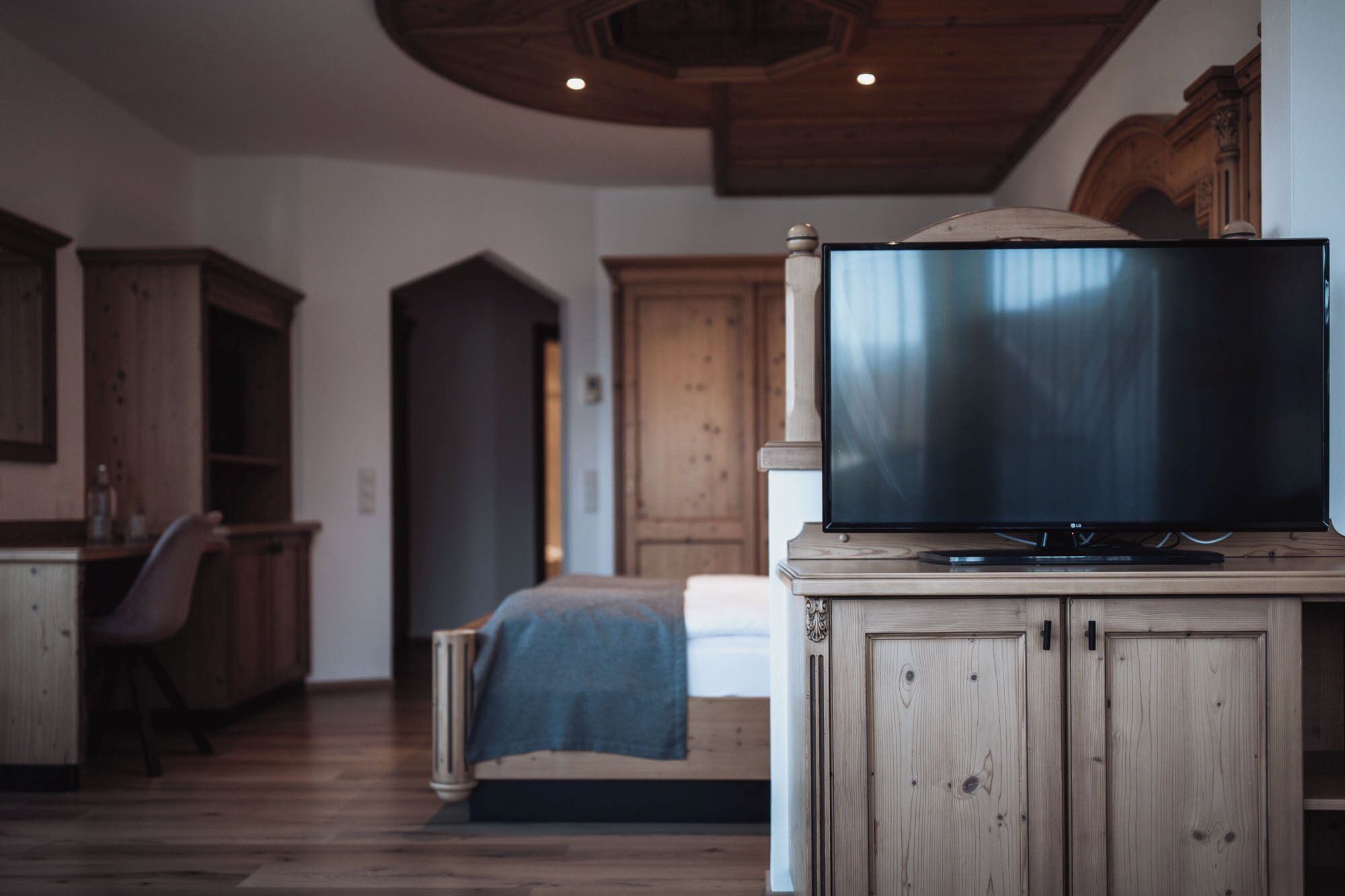 Quarto com cama, televisão sobre suporte de madeira, escrivaninha e cadeira.