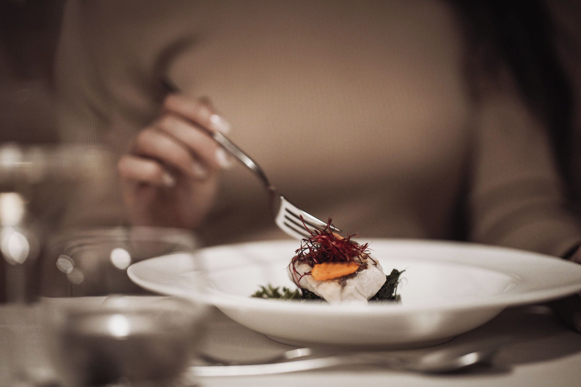 A person dining at a table with a white plate containing a dish, a fork, and a glass of water.