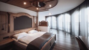 A bedroom with a large bed, a wooden headboard, a desk, and a large window with curtains. - Hotel Montis - Schönblick (Brunico)