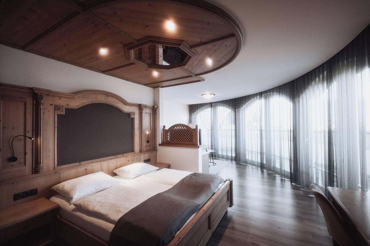 Une chambre à coucher avec un grand lit, une tête de lit en bois, un bureau et une grande fenêtre avec des rideaux.