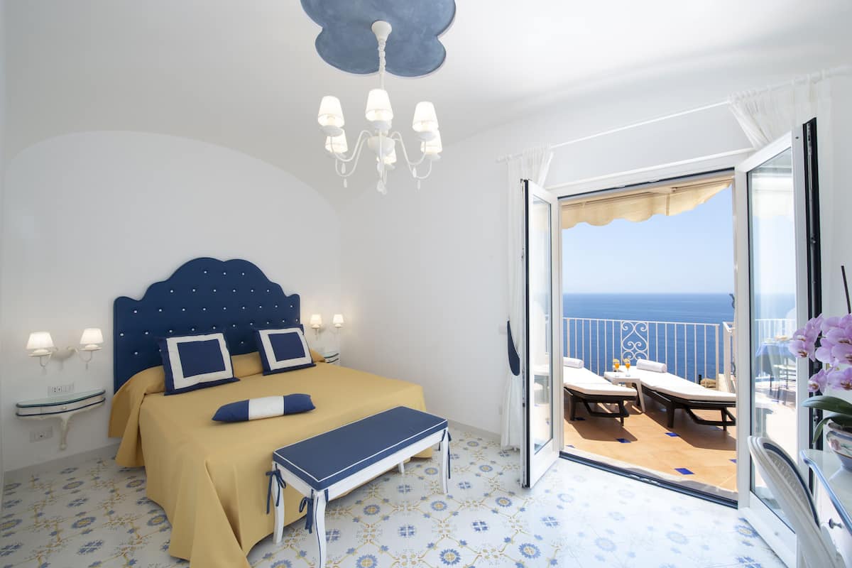 A bedroom with a bed, bedside lamps, a bench, and a view of the sea through an open door.