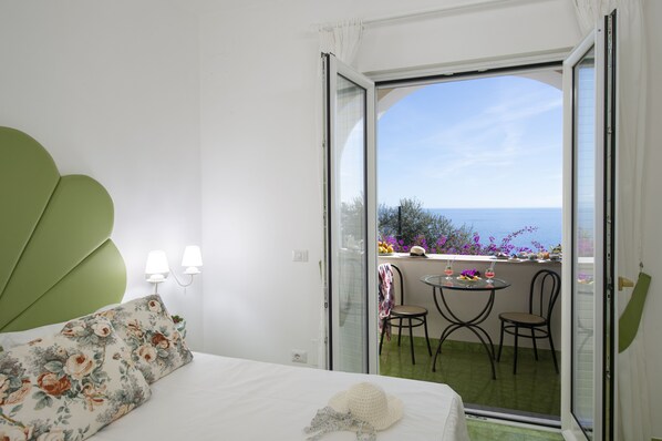Een slaapkamer met een bed, een groen hoofdeinde en een balkon met een tafel, stoelen en uitzicht op zee.