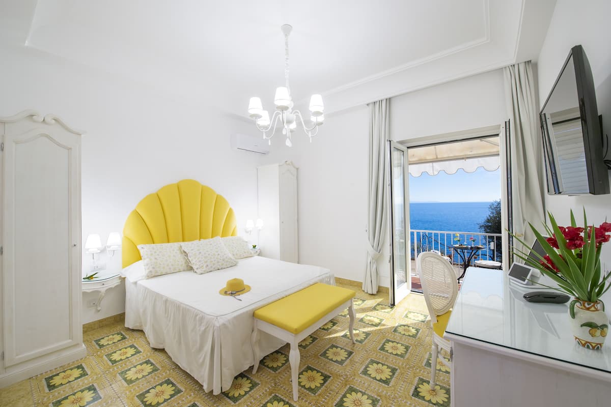A bright bedroom with a large bed, a small table, a bench, a TV, and a view of the sea.