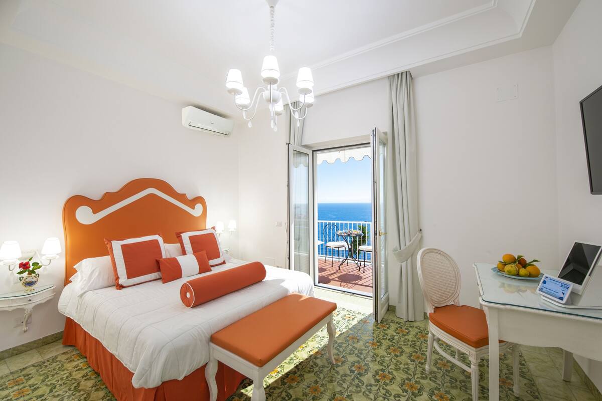 A hotel room with a bed, a desk with a laptop, a chair, a table with fruit, and a balcony with a view.