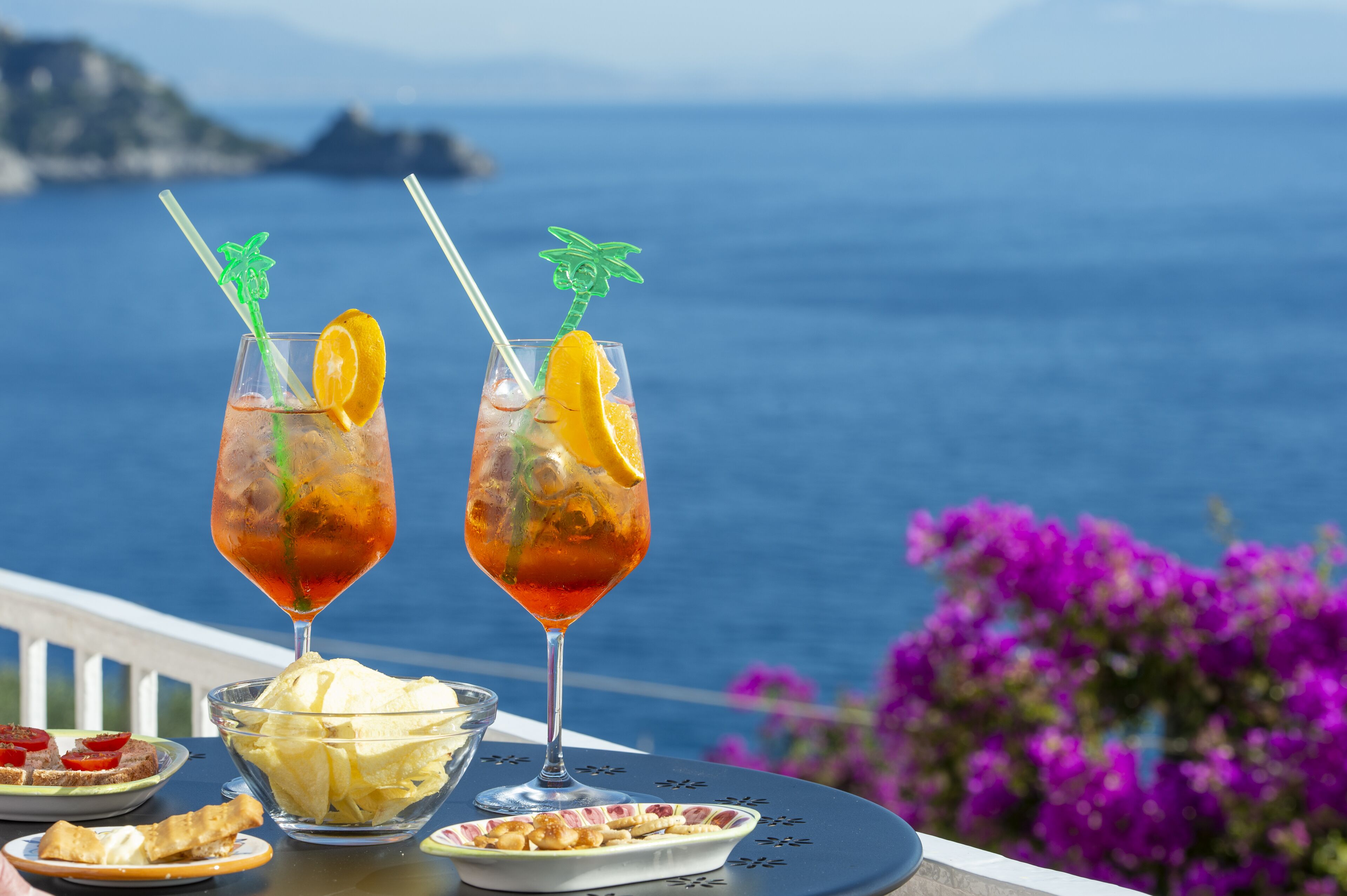 Two cocktails with garnishes on a table with a view of the sea.