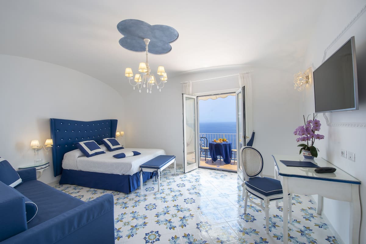 A bedroom with a bed, a desk, a chair, a TV, and a balcony with a view.