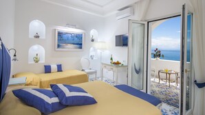 Habitación de hotel con dos camas, una mesita con una botella y fruta, una silla, un televisor y un balcón con vista al mar.