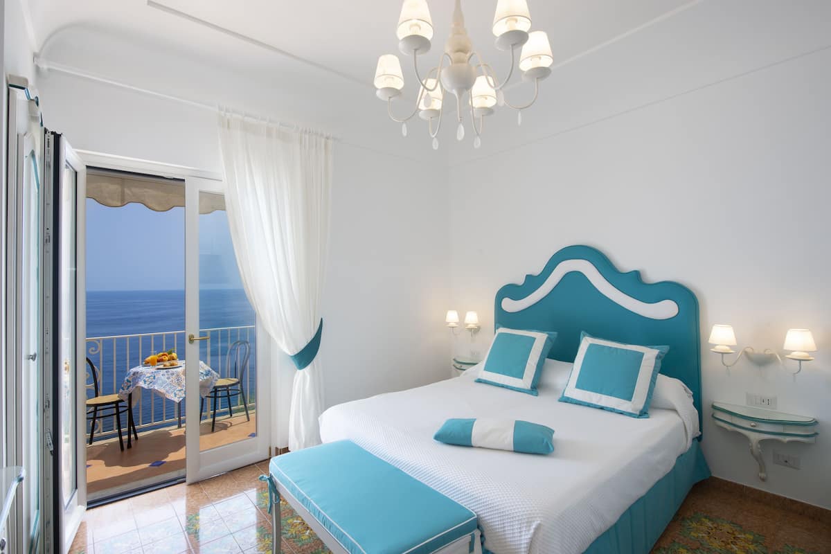 A bedroom with a bed, a nightstand, a chandelier, a window with curtains, and a balcony with a table and chairs.