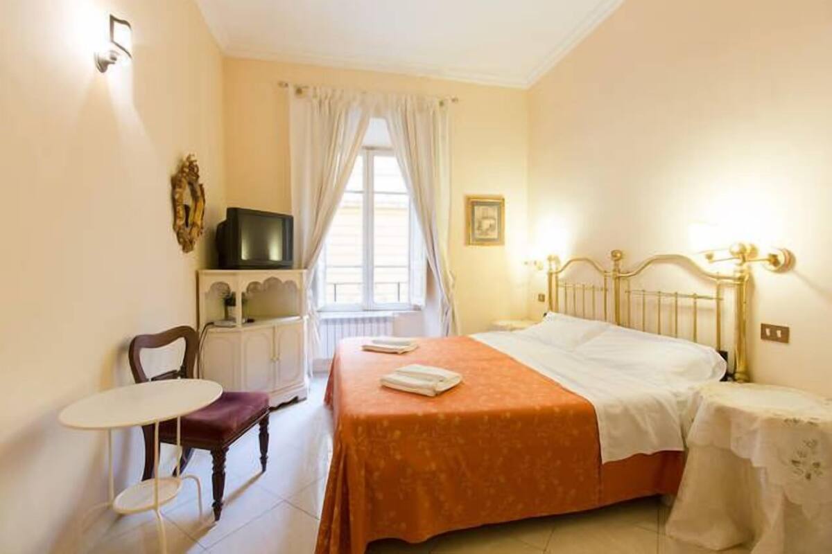 A bedroom with a bed, a chair, a small table, a television, and a window with curtains.