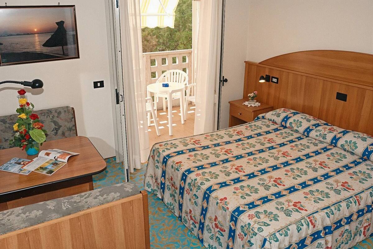 Une chambre d’hôtel avec un lit, un bureau et une chaise, une petite table avec un vase de fleurs, et une vue sur le balcon extérieur.