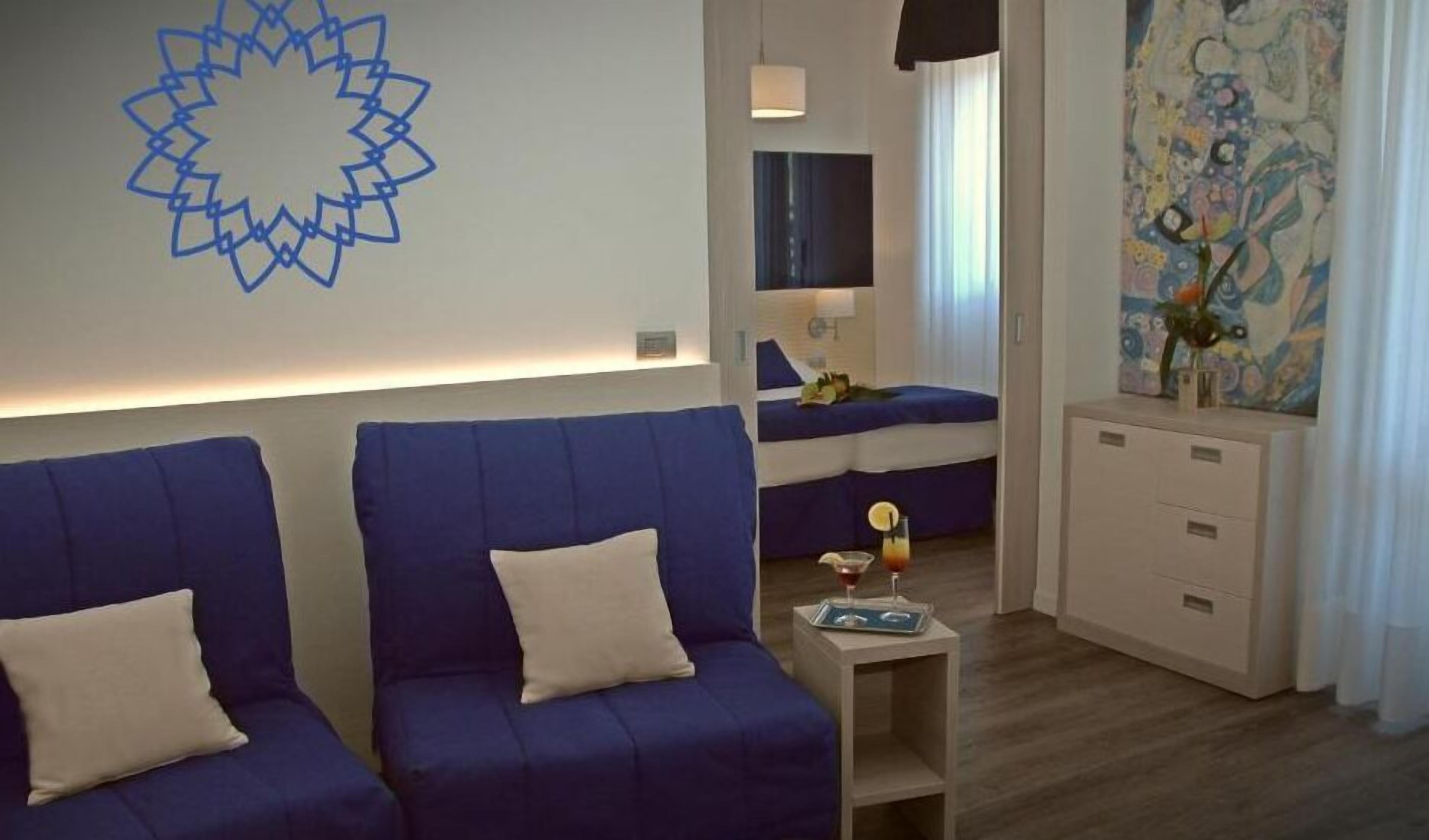 A modern hotel room with a blue sofa set, a bedside table with a glass of orange juice, and a colorful abstract painting on the wall.