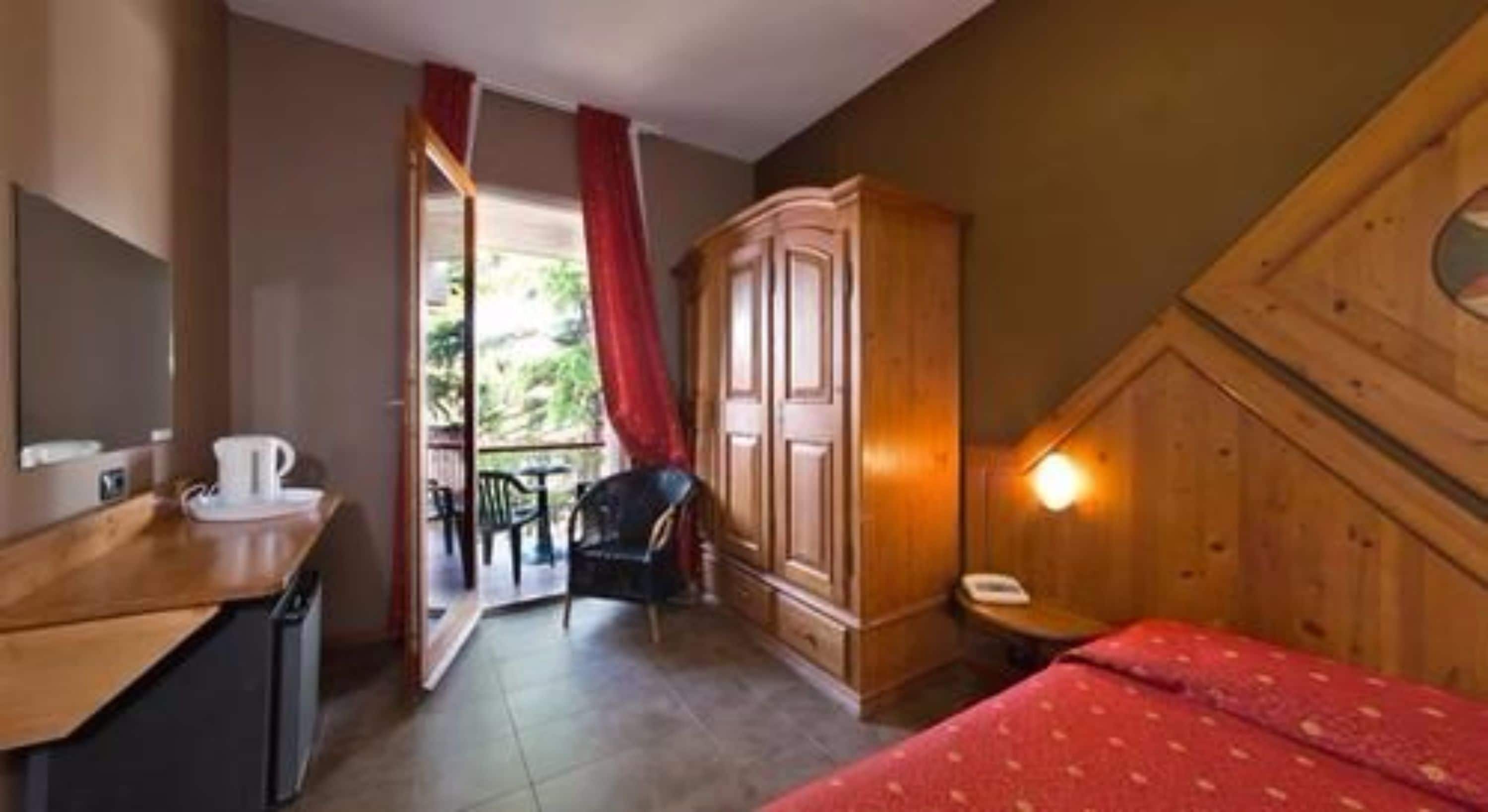 Um quarto pequeno e acolhedor com um guarda-roupa de madeira, uma escrivaninha com uma xícara, uma cadeira e uma cama com um cobertor vermelho.