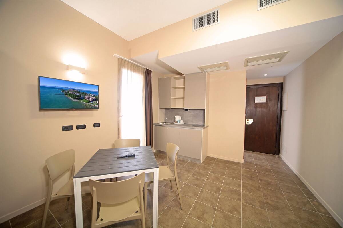 Een kleine kamer met een eettafel, stoelen, een aan de muur bevestigde televisie, een kitchenette en een deur.