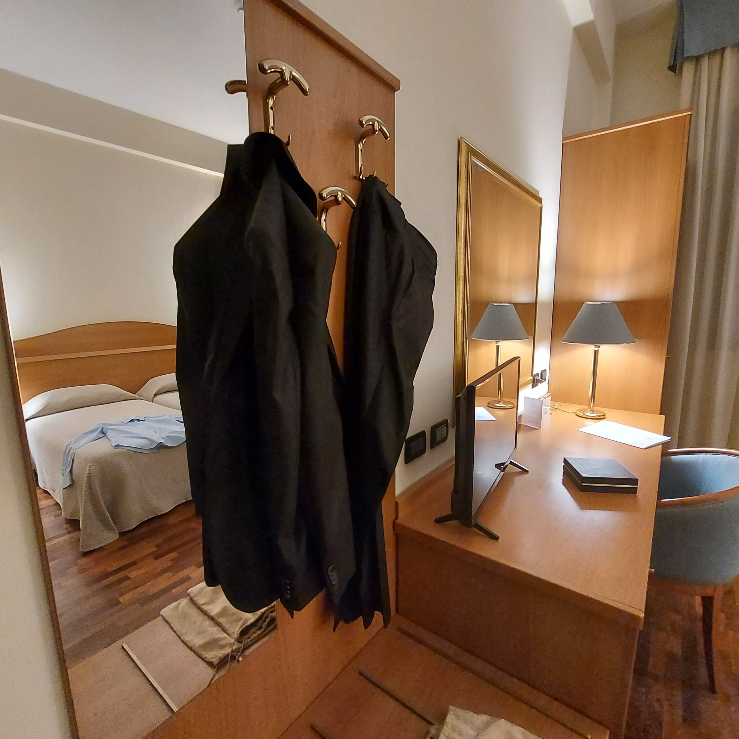 A hotel room with a bed, a desk with a lamp, a mirror, and two black jackets hanging on hooks.
