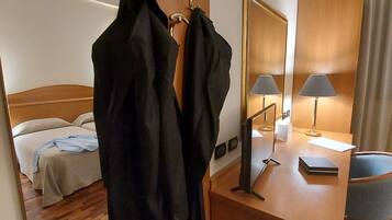 A hotel room with a bed, a desk with a lamp, a mirror, and two black jackets hanging on hooks.
