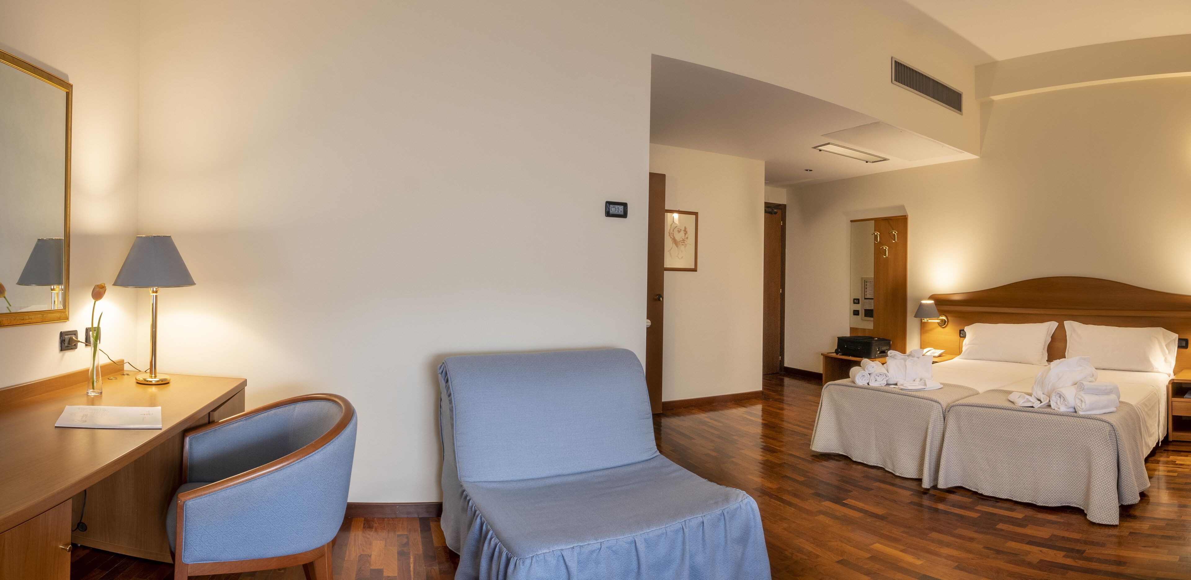 A hotel room with a wooden desk, a blue armchair, a bed with two bedside tables, and a large mirror.