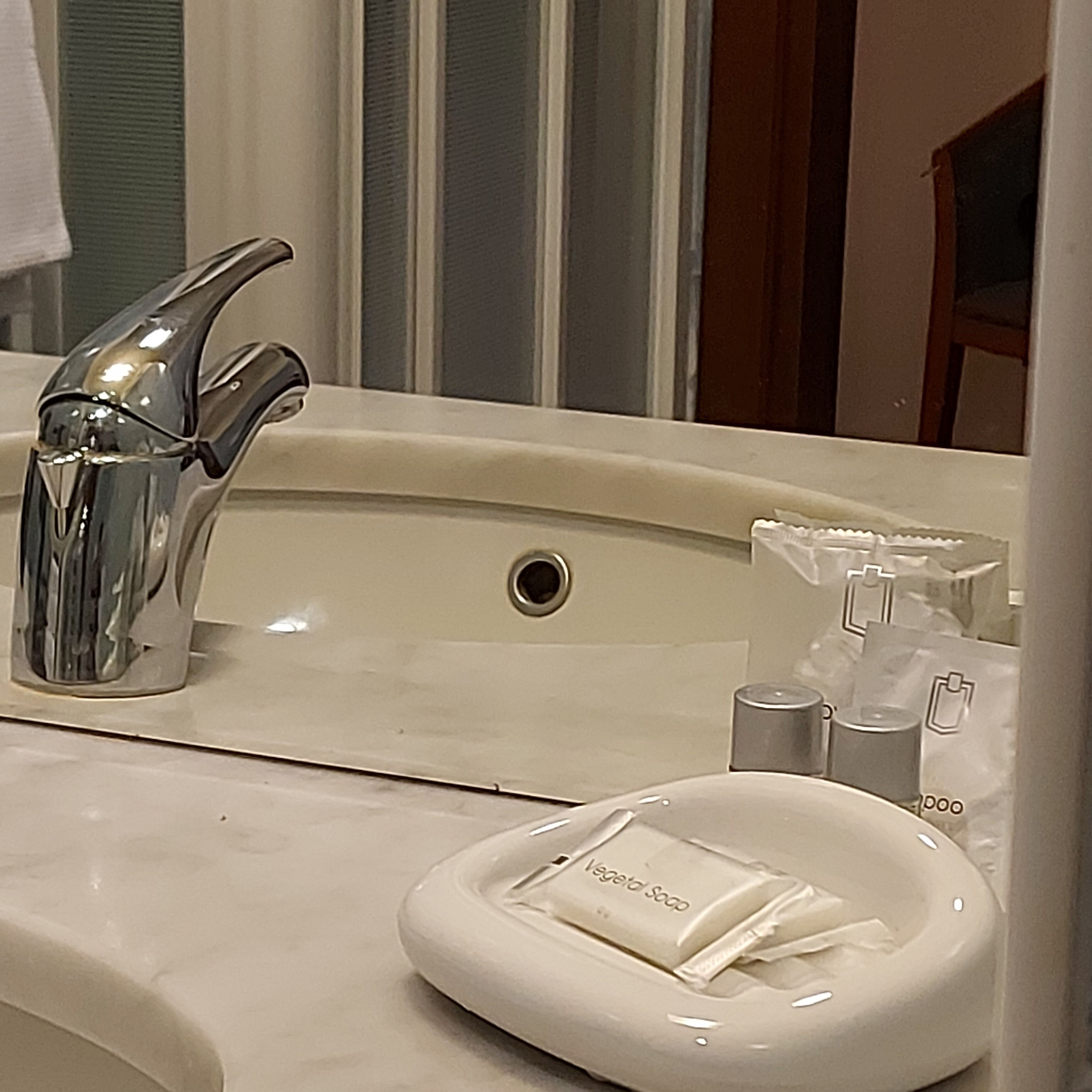 Ein Badezimmerwaschbecken mit chromfarbenem Wasserhahn, einem Spiegel und Toilettenartikeln, darunter eine Seifenschale.