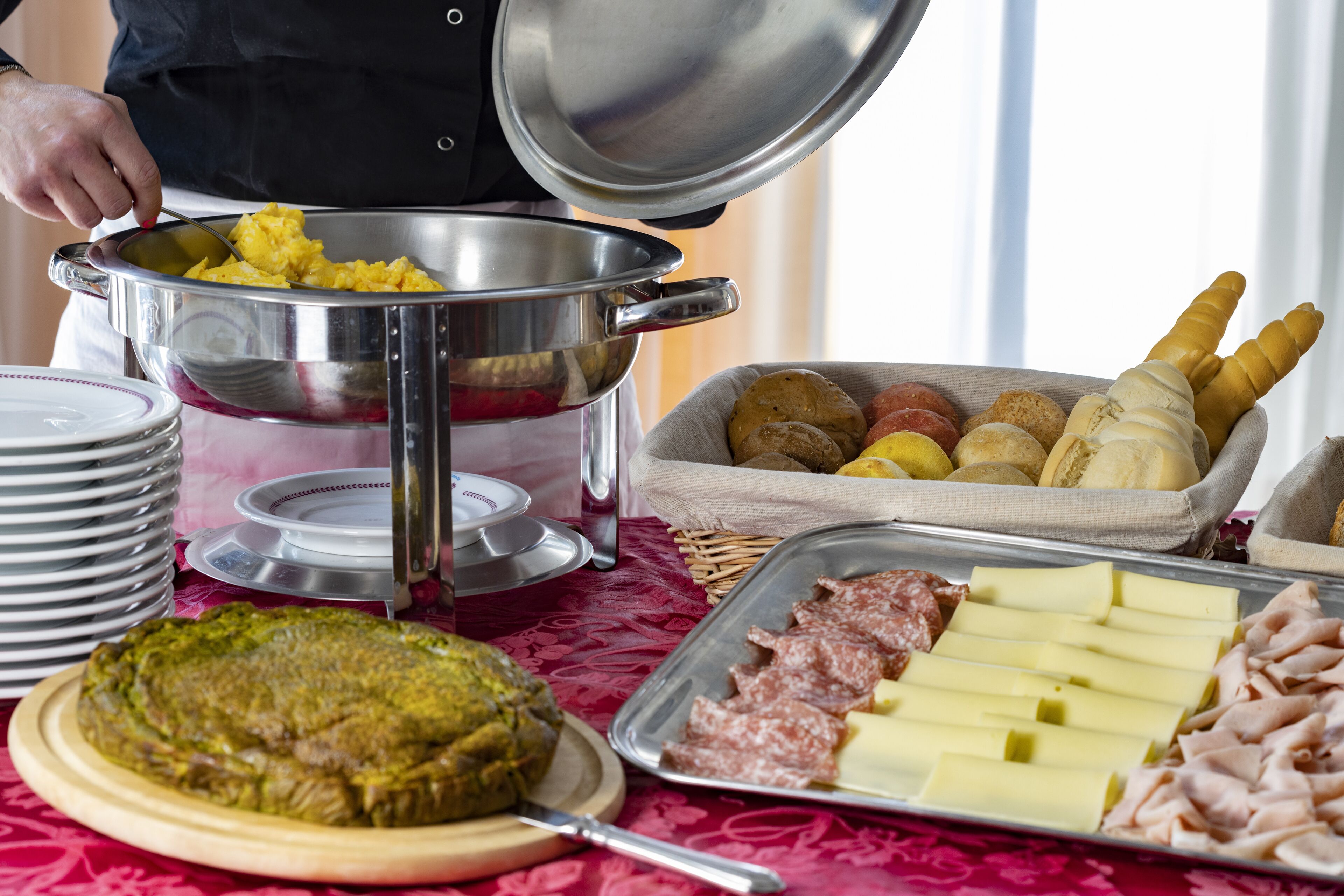 Une table de buffet proposant divers plats, notamment un plat d’œufs brouillés, un plateau de viandes et un saladier de pain.