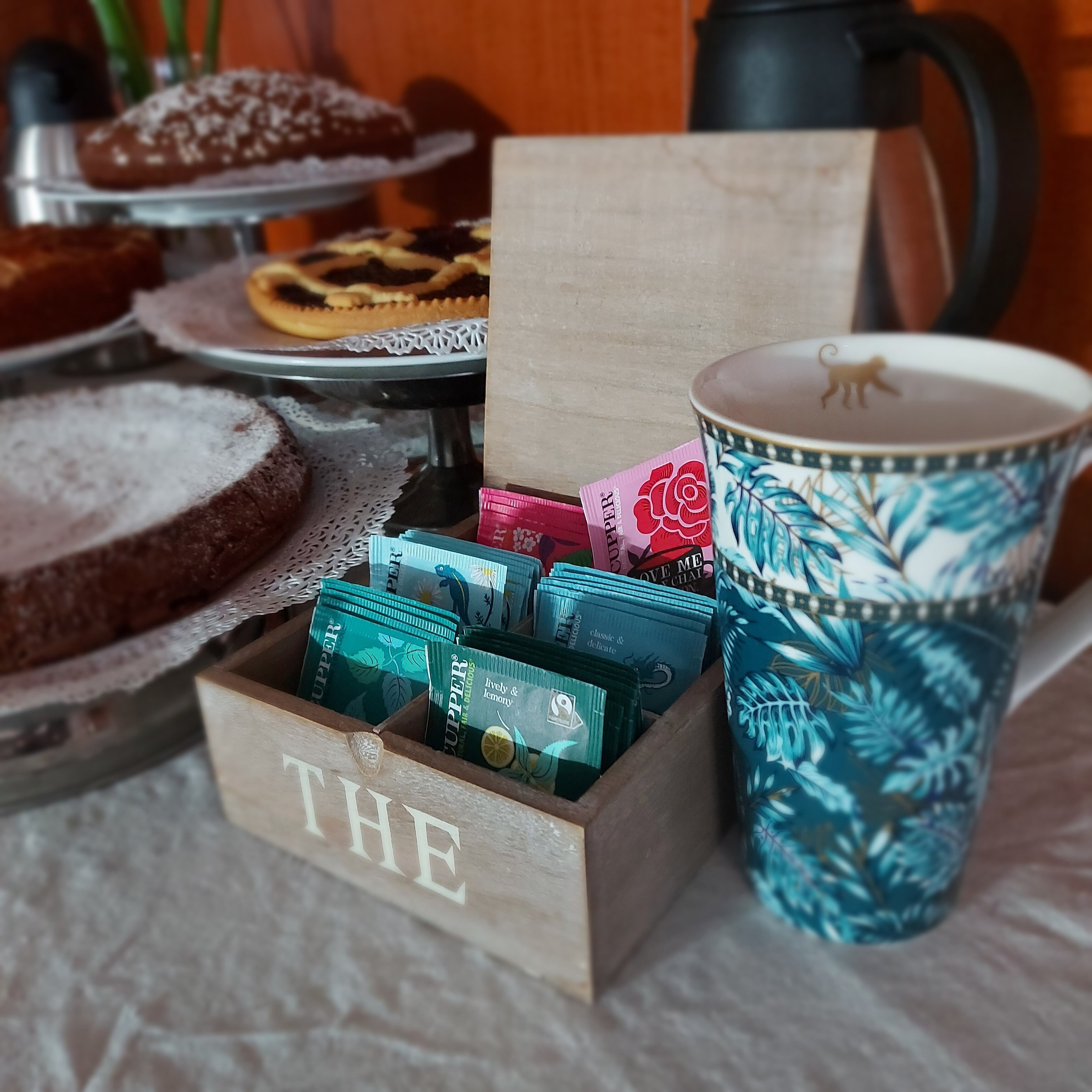 A wooden box labeled "THE" filled with tea tins, a patterned mug, and a tray with pastries.