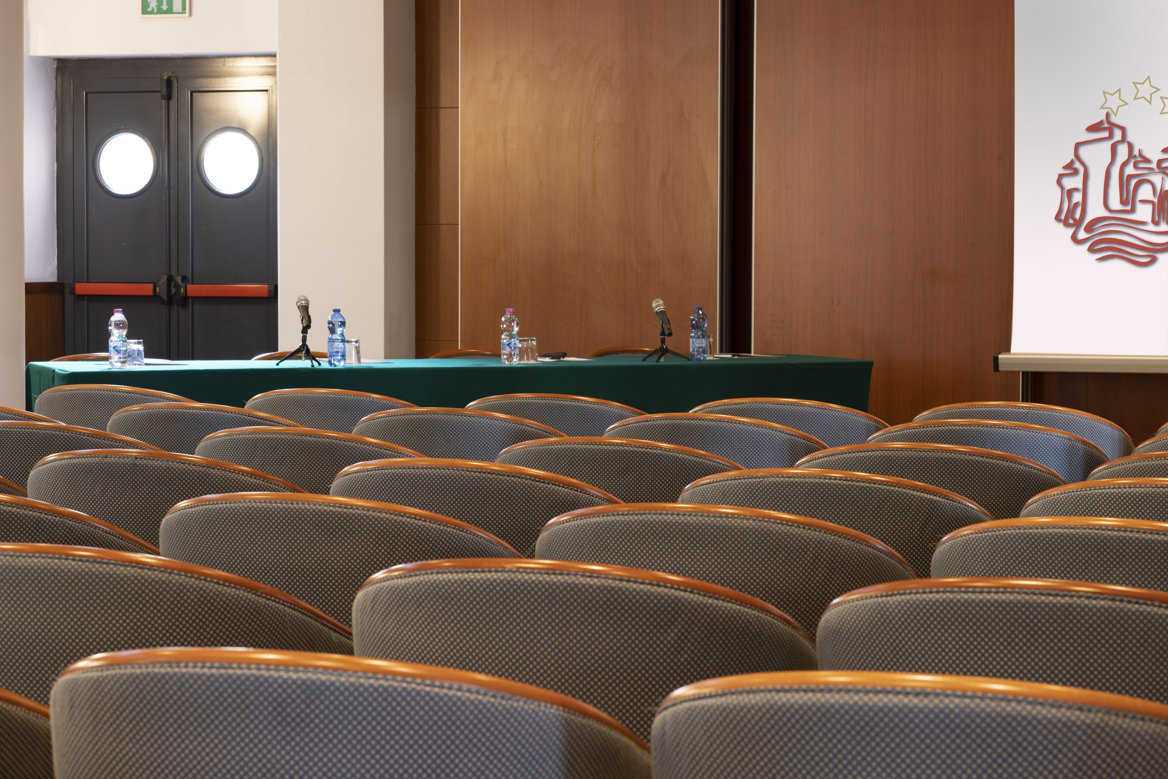 Une salle de conférence avec des rangées de chaises, une table équipée de microphones, des bouteilles d’eau et une porte en bois ornée de fenêtres rondes.