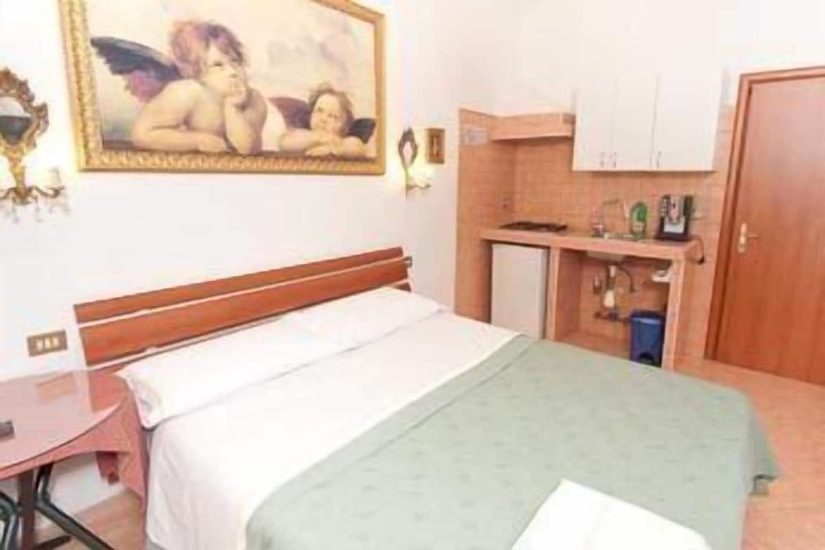 A hotel room with a bed, a small desk, a kitchenette, and a painting on the wall.