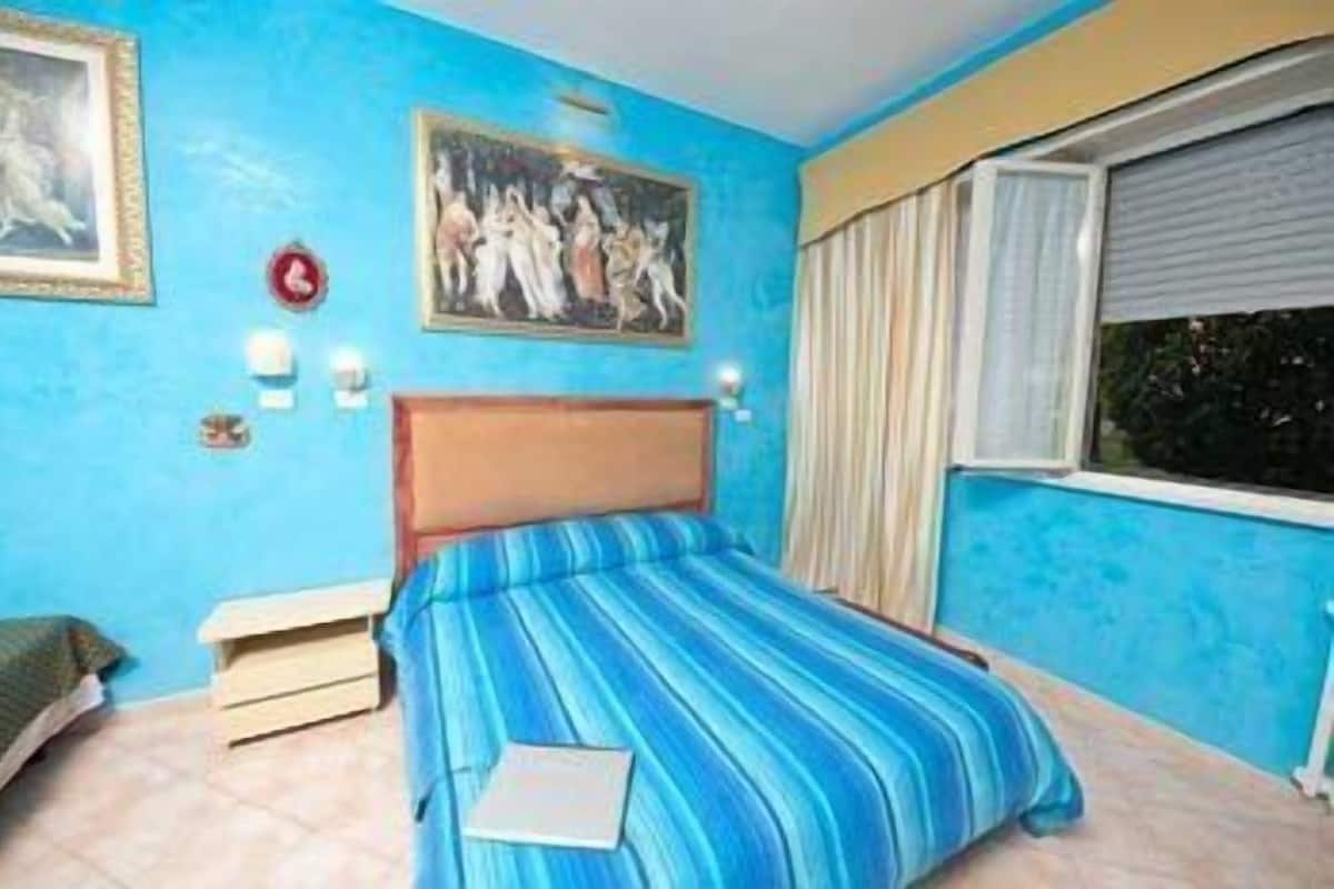 A bedroom with a bed, a nightstand, a painting, a window with curtains, and a wall clock.