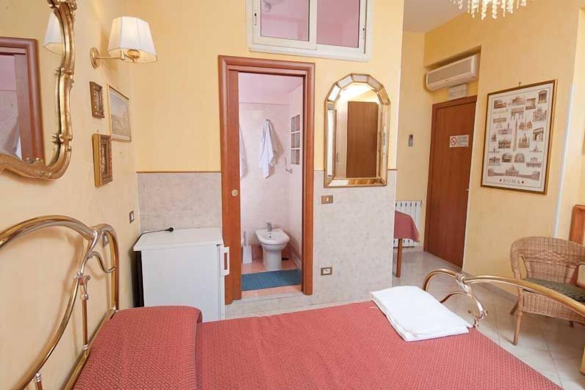 A hotel room with a bed, a small desk, a chair, a mirror, and a door leading to a bathroom.