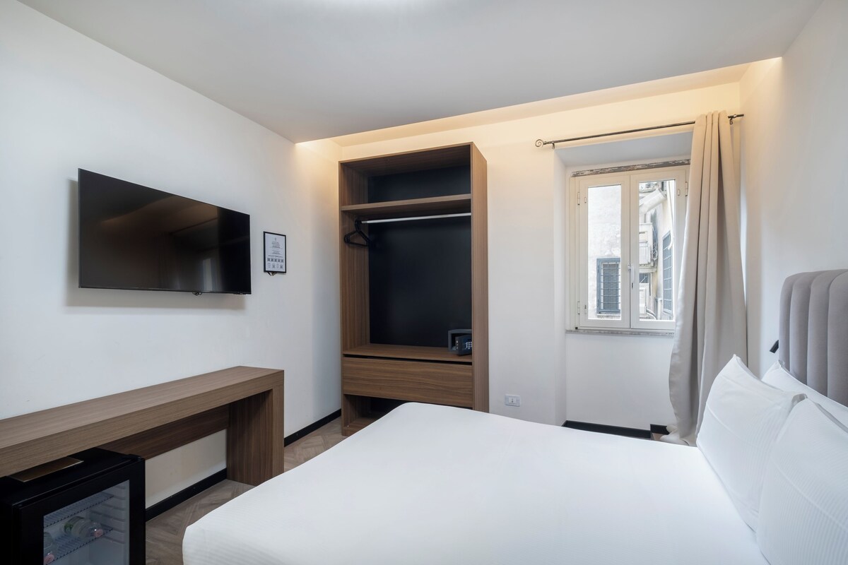 A hotel room with a bed, a TV mounted on the wall, a wooden wardrobe, a bench, and a window with curtains.