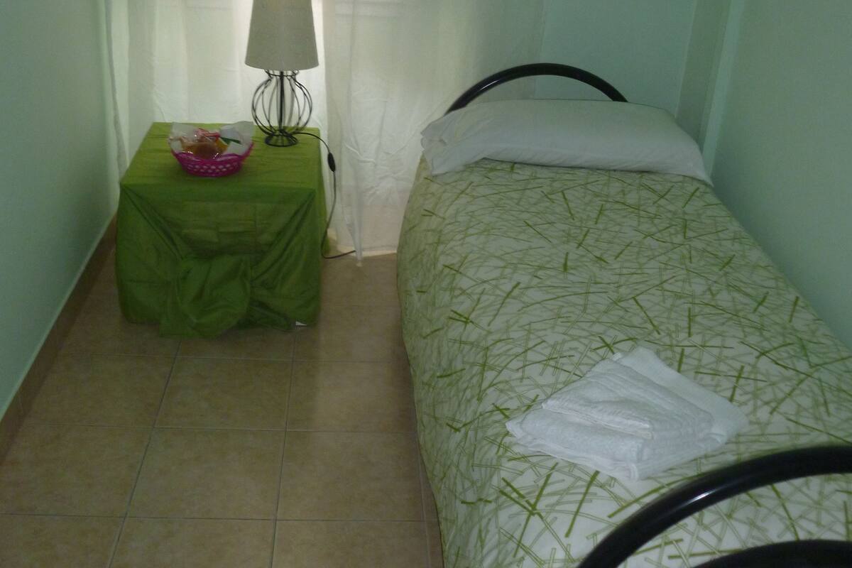 A small, single-bed room with a green bedspread, a bedside table with a lamp, and a curtain partially drawn.