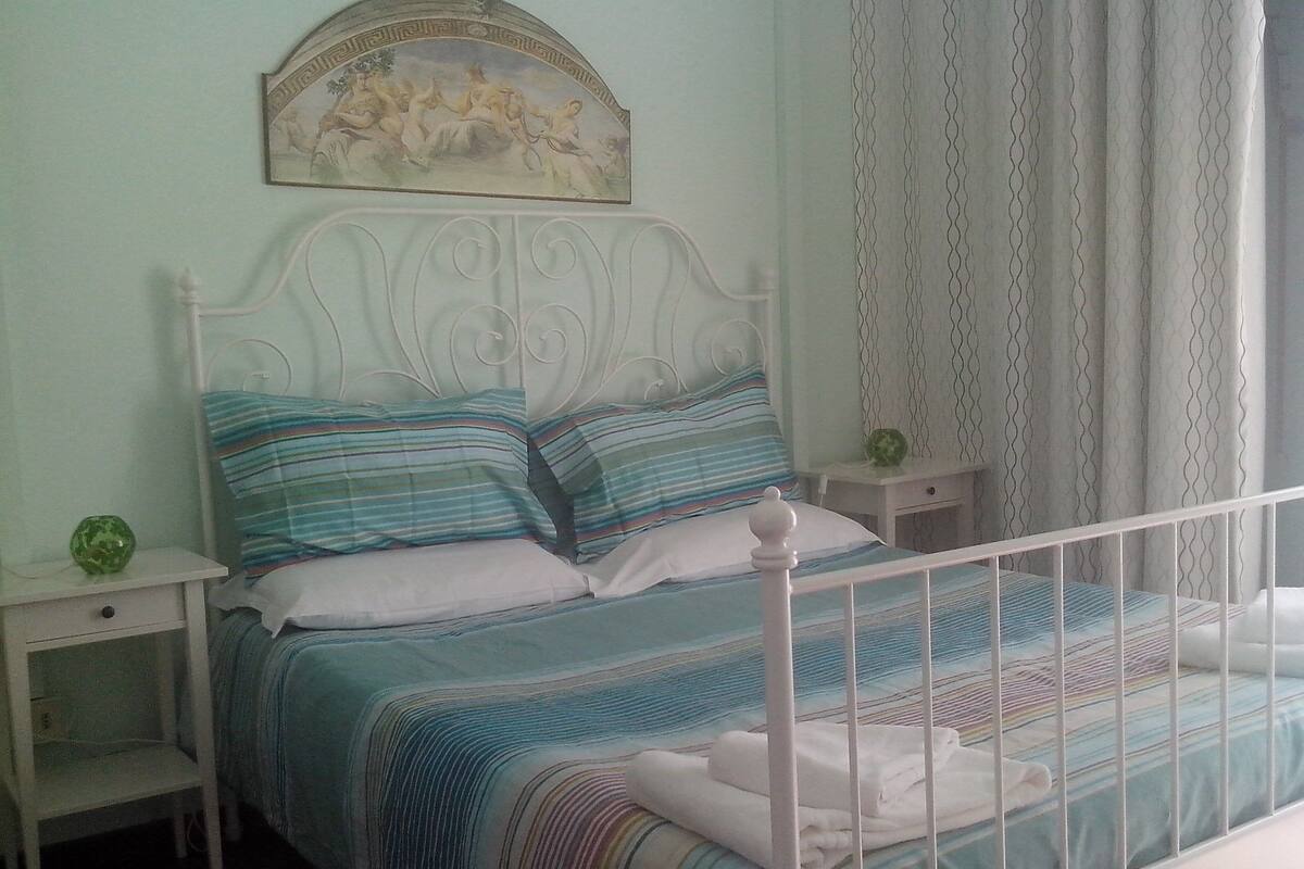 A bed with a white metal frame, a blue and white striped bedspread, and two pillows. There is a small bedside table with a green object on it. A painting is above the bed, and a curtain is partially visible on the right.