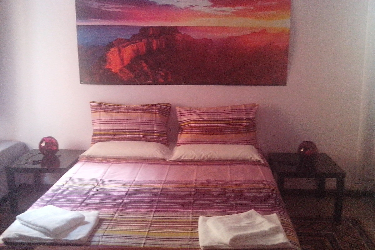 A hotel room with a bed, two pillows, a nightstand, a lamp, and a painting of a sunset.