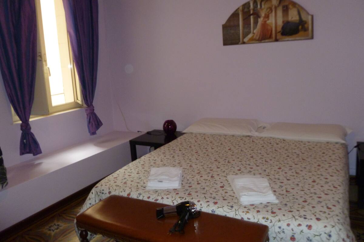 A bedroom with a bed, a bench, a small table, a painting on the wall, and a window with curtains.