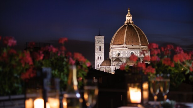 Uma mesa de jantar com velas e flores, vista para um edifício com cúpula e torre, e céu noturno.