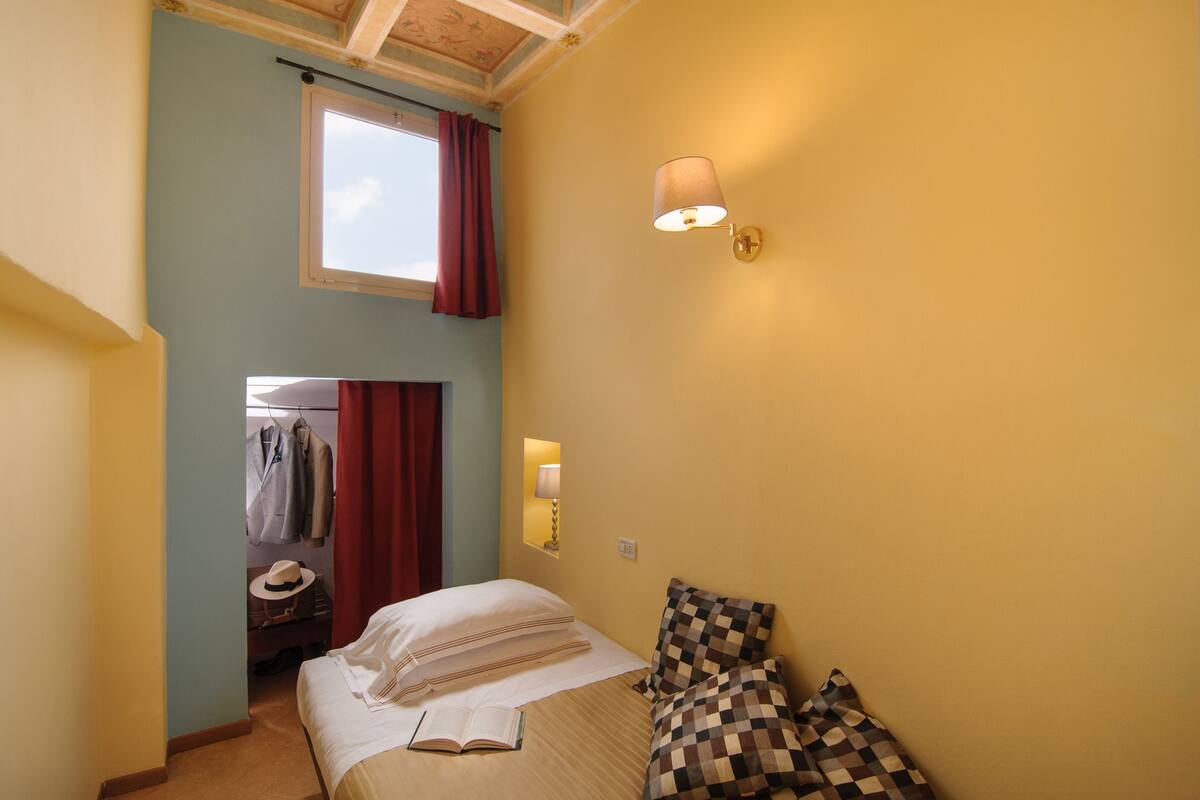 Ein Schlafzimmer mit Bett, Kissen, Nachttisch, Lampe, einem Fenster mit Vorhängen und einem Spiegel.